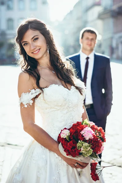 Happy brunette bride. She is holding the wedding bouquet of red and pink flowers at the background of the standing groom. The street portrait. — Stock Photo, Image