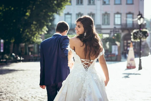 Tillbaka med utsikt över den bedårande leende brunett bruden med bar rygg. Brudgummen leder henne med handen längs den soliga staden gatan. — Stockfoto