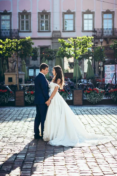 Charmant lächelnde Frischvermählte in eleganter Kleidung halten zärtlich Händchen auf der sonnigen Straße. Hochzeitsporträt in voller Länge. — Stockfoto