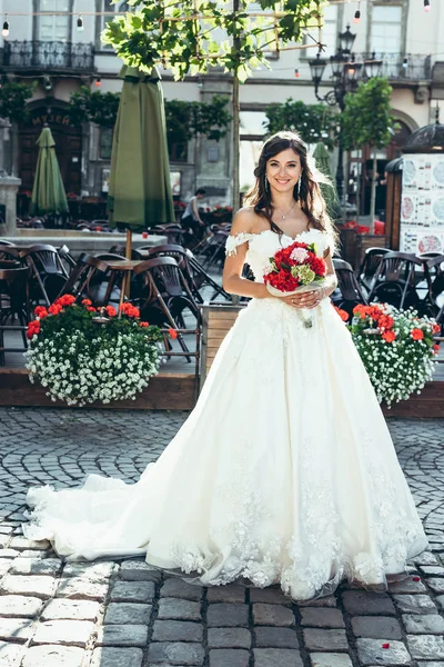 Retrato de comprimento total da noiva morena sorridente no vestido de casamento longo segurando o buquê de flores vermelhas e rosa na rua coberto flores wtih . — Fotografia de Stock
