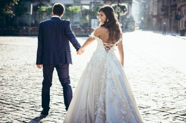 Gülümseyen güzel esmer gelin elbise içinde güneşli Caddesi boyunca çıplak sırt ile önde gelen damadın arkadan görünüm.