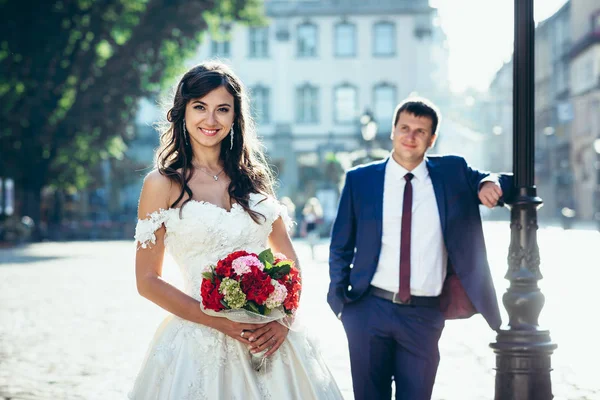 Usměvavá nevěsta s červené a růžové svatební kytici květin na rozostřeného pozadí ženicha, opírající se o pouliční lampu. — Stock fotografie