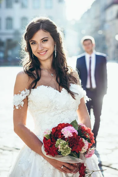 Usmívající se bruneta nevěsta s přírodní make-up svatební kytice červené a růžové květy v drží rozostřeného pozadí ženicha. Venkovní portrét. — Stock fotografie