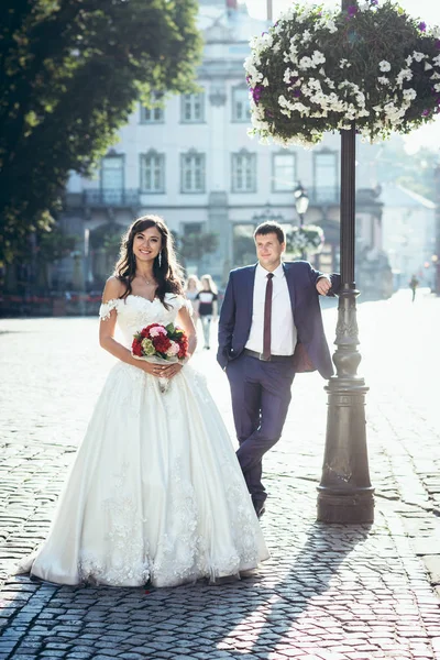 Retrato de casamento ao ar livre completo. A bela noiva morena com o buquê de flores vermelhas e rosa está de pé no fundo do noivo inclinado sobre a lâmpada de rua . — Fotografia de Stock