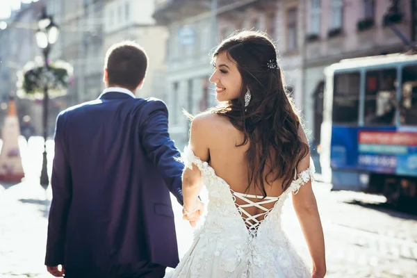 Sposo sta conducendo per mano la sua adorabile sposa bruna sorridente nel vestito con la schiena nuda lungo la strada della città. Il ritratto posteriore . — Foto Stock