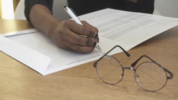 A rapariga na recepção está a preencher a informação no formulário. A vista de perto das mãos africanas. Os óculos deitados sobre a mesa . — Vídeo de Stock