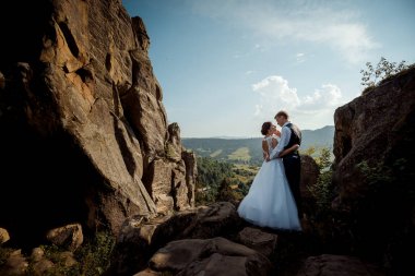 Yakışıklı sarışın damat yavaşça büyüleyici gelini güneşli kayaların üzerinde dururken yanak dokunuyor.