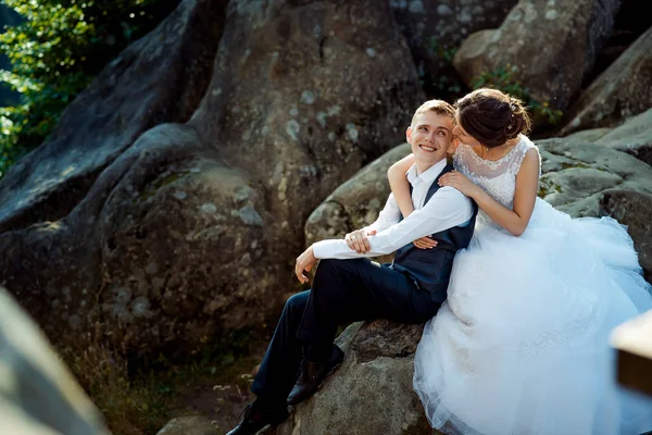 Abraço sorridente casal recém-casado estão olhando uns para os outros enquanto sentado na rocha. Retrato de casamento close-up . — Fotografia de Stock