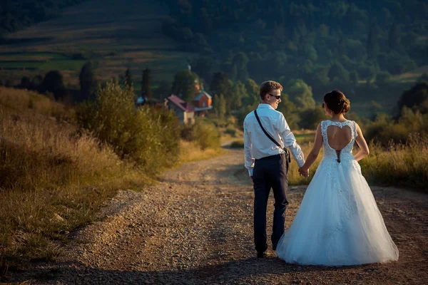 Visão traseira dos recém-casados de mãos dadas e caminhando ao longo da estrada rústica para as montanhas durante o pôr do sol . — Fotografia de Stock