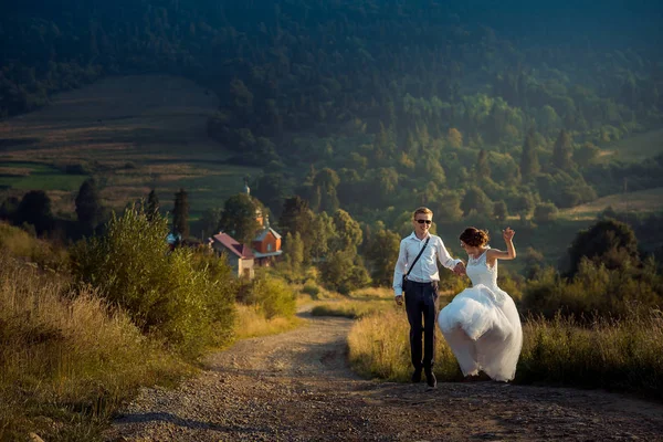 Весільний знімок радісного привабливого щойно одружився в сонцезахисних окулярах, танцюючи o nthe Road на тлі гір під час заходу сонця . — стокове фото