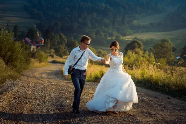 Encantador jovem feliz casal recém-casado está rindo e de mãos dadas durante a sua caminhada ao longo do campo durante o pôr do sol teh . — Fotografia de Stock