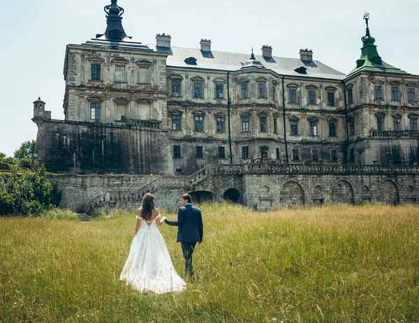 Ganzheitliche Rückansicht des stilvollen Brautpaares, das sich Händchen hält und bei sonnigem Wetter zum alten Schloss spaziert. — Stockfoto