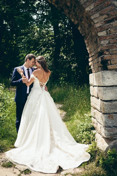 Retrato al aire libre de larga duración de los recién casados felices y atractivos abrazando suavemente y frotando narices bajo el arco de piedra en el bosque . — Foto de Stock