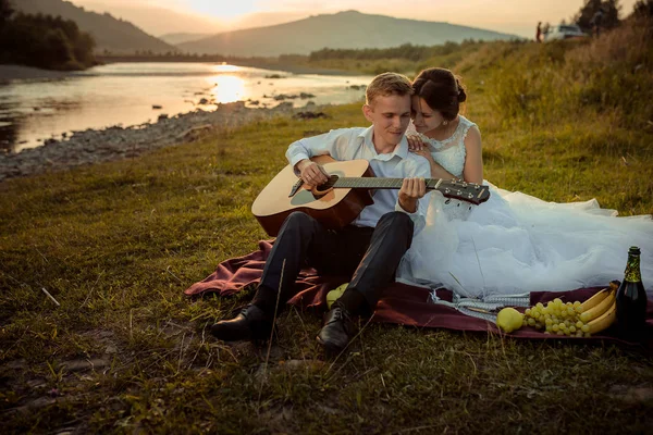 Romantisches Hochzeitsporträt während des Sonnenuntergangs. schöner Bräutigam spielt Gitarre und die hinreißende Braut umarmt ihn während ihres Picknicks am Fluss auf dem Plaid sitzend. — Stockfoto
