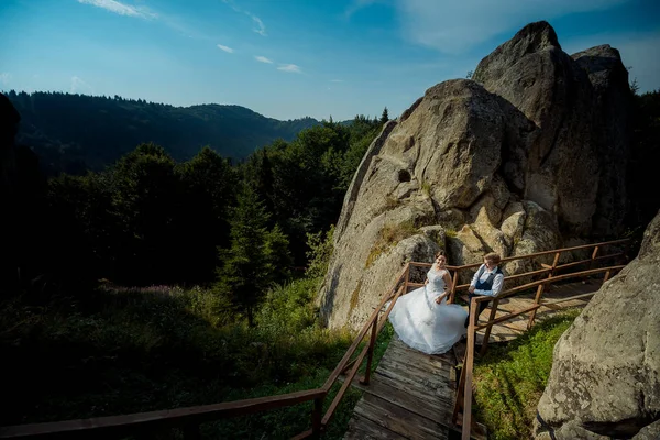 Noiva sorridente no vestido de casamento longo está girando ao redor perto do noivo nas escadas de madeira nas belas montanhas . — Fotografia de Stock