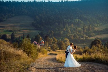 Neşeli şık yeni evliler günbatımı sırasında kırsalında yolda şefkatle sarılıyorlar. Tam uzunlukta açık atış.