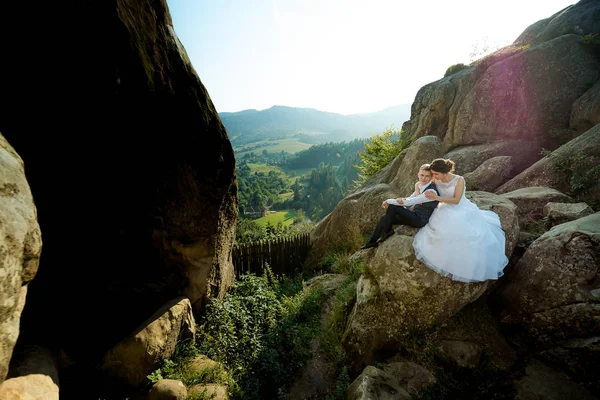 Чувствительный свадебный портрет на открытом воздухе. Невеста обнимает жениха, сидя на скале на фоне прекрасного солнечного пейзажа . — стоковое фото