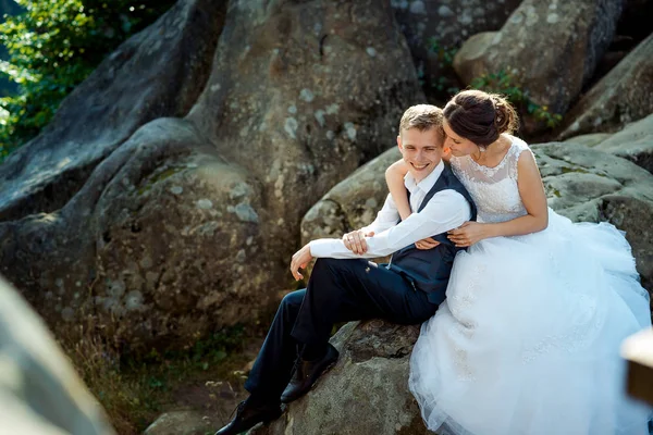 Atractivos jóvenes recién casados se abrazan tiernamente mientras están sentados en la roca . — Foto de Stock