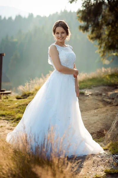 Bonita novia joven con el pelo hacia arriba, maquillaje natural y sonrisa encantadora posando en el bosque durante la puesta de sol. Boda de cuerpo entero . — Foto de Stock