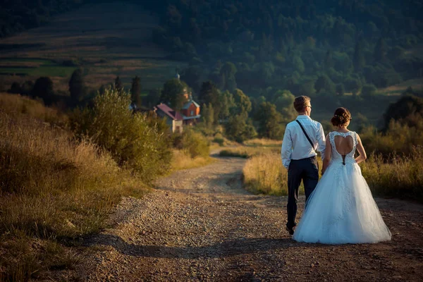 Gladlynt promenader nygift par längs vägen till de magnifika berg under solnedgången. Baksidan Bröllop skott. Par håller händerna. — Stockfoto