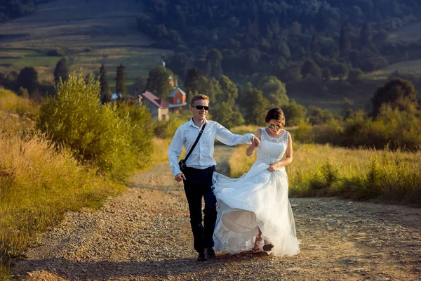 Vue de face du beau jeune couple jeune marié tenant la main, souriant et marchant activement le long de la route dans la campagne pendant le coucher du soleil . — Photo