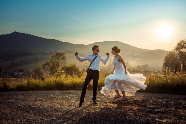 Attraktiva lyckligt nygifta har roligt medan du dansar på vägen på landsbygden under solnedgången. — Stockfoto