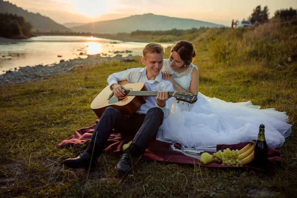 Composizione del picnic nuziale sulla riva del fiume durante il tramonto. Sorridente sposo sta giocando la chitarra, mentre la sposa lo sta abbracciando indietro mentre seduto sul plaid . — Foto Stock