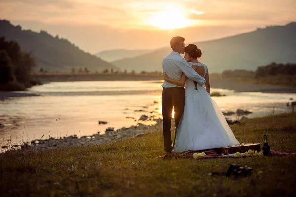 Groom está beijando suavemente sua linda noiva na testa durante o pôr do sol. Piquenique de casamento na margem do rio. Bela paisagem . — Fotografia de Stock