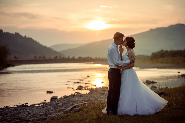 Adorable pareja recién casada se besa suavemente en la orilla del río durante la puesta del sol . —  Fotos de Stock