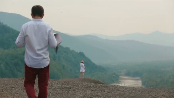 La vue arrière du touriste tenant la carte et regardant la belle femme blonde debout sur le bord des montagnes. Vue sur drone . — Video