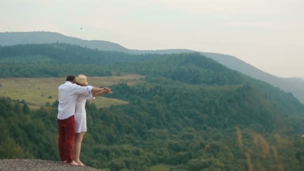 O homem está segurando a mão e abraçando a linda garota loira de volta enquanto está na borda das montanhas e desfrutando da paisagem . — Vídeo de Stock