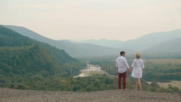 Вид сзади с дрона на счастливую влюбленную пару, держащуюся за руки, стоя на краю гор и наслаждаясь пейзажем. Мужчина целует ее в щеку . — стоковое видео