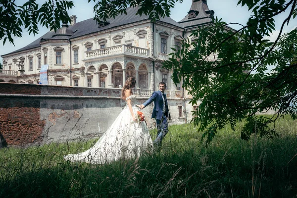 L'elegante coppia di sposi si tiene per mano mentre cammina lungo la foresta verde sullo sfondo della vecchia casa . — Foto Stock