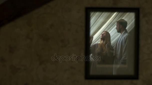 El reflejo en el espejo de la hermosa rubia en la bata mirando por la ventana mientras su novio la abraza suavemente la espalda . — Vídeo de stock