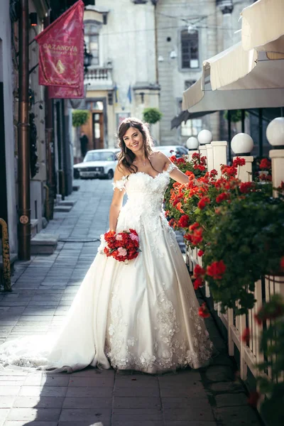 Den charmiga brunett bruden med vackra leende håller bröllop bukett av röda och rosa blommor i soliga gatan. Fullängds utomhus porträtt. — Stockfoto