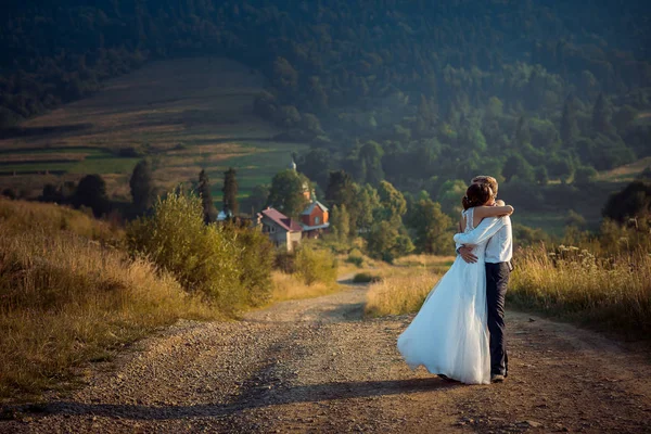 Casal recém-casado elegante está abraçando na estrada no fundo das belas montanhas durante o pôr do sol . — Fotografia de Stock