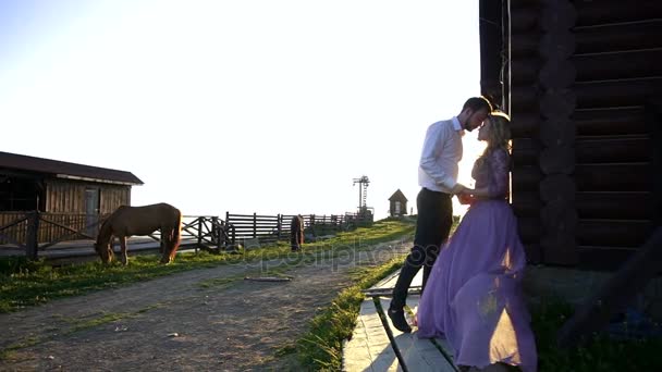 Bonito homem está de mãos dadas, tocando narizes e beijando com sua encantadora namorada loira no vestido roxo longo ao sol. Eles estão se apoiando na velha casa de madeira no campo . — Vídeo de Stock