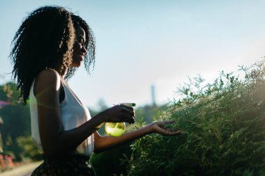 Doğal makyaj ve güzel gülümseme için yeşil bitkiler yeşil plastik tragger sprey kullanarak güzel büyüleyici Afrikalı kız portresi yan.