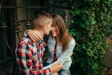 Genç sevgili çift yeşil bitkilerle kaplı duvarın yanında otururken gülümseyen sarılma.