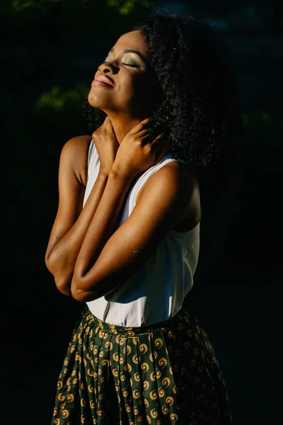 Retrato de meio comprimento da encantadora menina africana sorridente com sombras de olhos verdes desfrutando da luz do sol no jardim . — Fotografia de Stock
