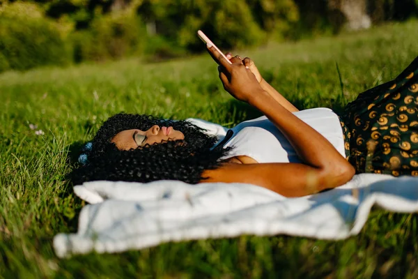 Straně portrét mladé atraktivní Africká dívka s zelené oční stíny, kterým se na trávě a procházení a chatování přes mobilní telefon. — Stock fotografie