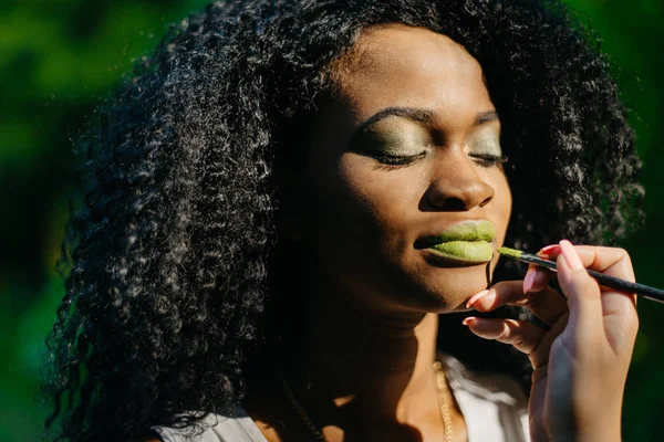 Mão de maquiador profissional aplicando o batom verde usando escova no rosto de menina afro-americana encantadora com sombras de olhos verdes no fundo verde . — Fotografia de Stock