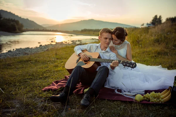 Привлекательный жених играет на гитаре, и его очаровательная невеста наслаждается этим во время их пикника на берегу реки во время заката . — стоковое фото