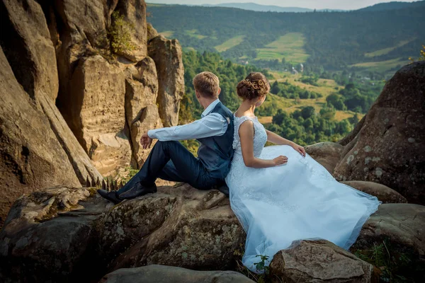 Glamour newlywed para siedzi na skale back-to-back i widoki krajobraz w słoneczny dzień. — Zdjęcie stockowe