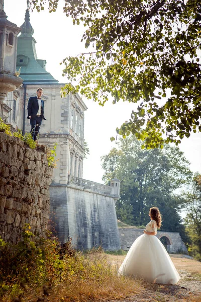 Ganzheitliches Hochzeitsporträt der Braut, die den Bräutigam auf der Steinmauer vor dem Hintergrund des alten Schlosses betrachtet. — Stockfoto