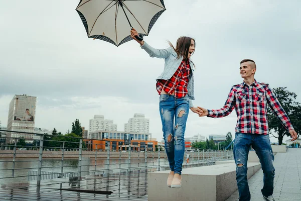 Hermosa chica alegre está saltando con paraguas y la mano de su novio sonriente mientras camina a lo largo de Ostrow Tomski en Wroclaw, Polonia . — Foto de Stock