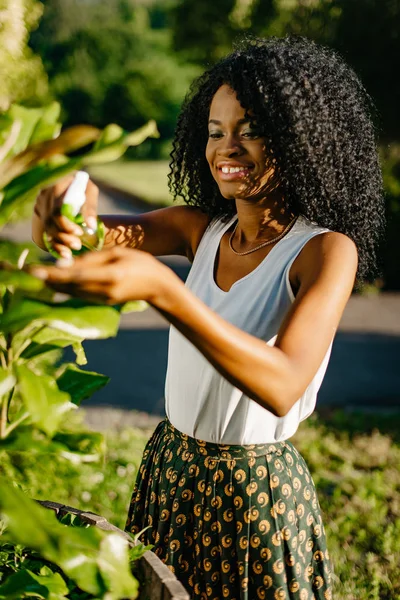 Вертикальный солнечный портрет. Красивая молодая африканская девушка с очаровательной улыбкой и зелеными тенями для век с помощью зеленого пластикового триггера спрей для наружных растений . — стоковое фото