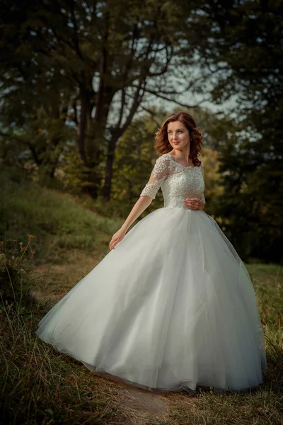 Full-length wedding portrait of the beautiful ginger hair bride in the long wedding dress with red lipstick in the forest.