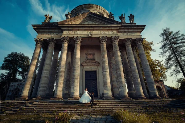 Ženich je drží ruku okouzlující nevěstu a stojící na jedno koleno vedle vlasy na schodech starého barokního kostela. — Stock fotografie