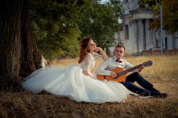 Affascinante sposa sorridente sta ascoltando lo sposo elegante suonare la chitarra mentre seduto sull'erba sotto l'albero . — Foto Stock